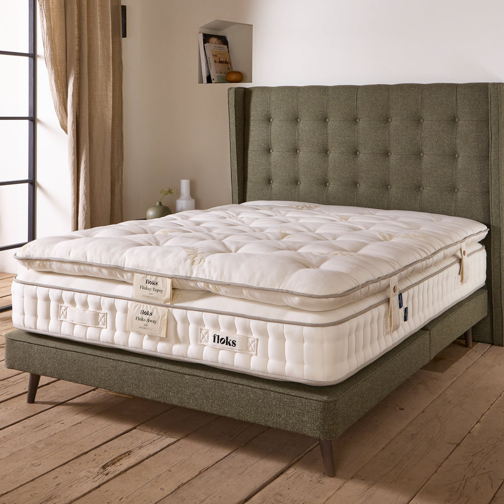 Wide image of a Floksy Topsy mattress enhancer on top of a Floks Away mattress on a wooden floor and green upholstered 'Thomas' bed.