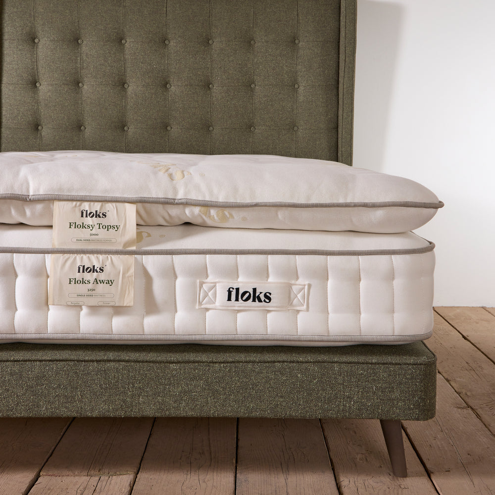 Close up of a Floksy Topsy mattress enhancer on a wooden floor and green upholstered headboard.